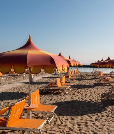 Spiaggia con ombrelloni arancioni e lettini su sabbia, cielo sereno.