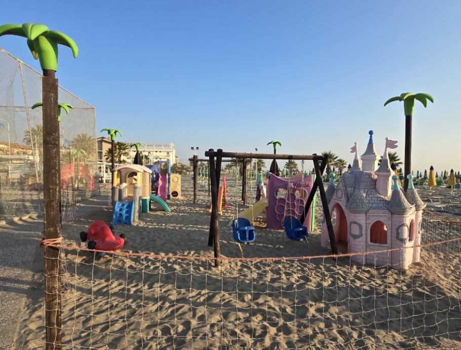 Area giochi sulla spiaggia con altalene e castello di plastica colorato.