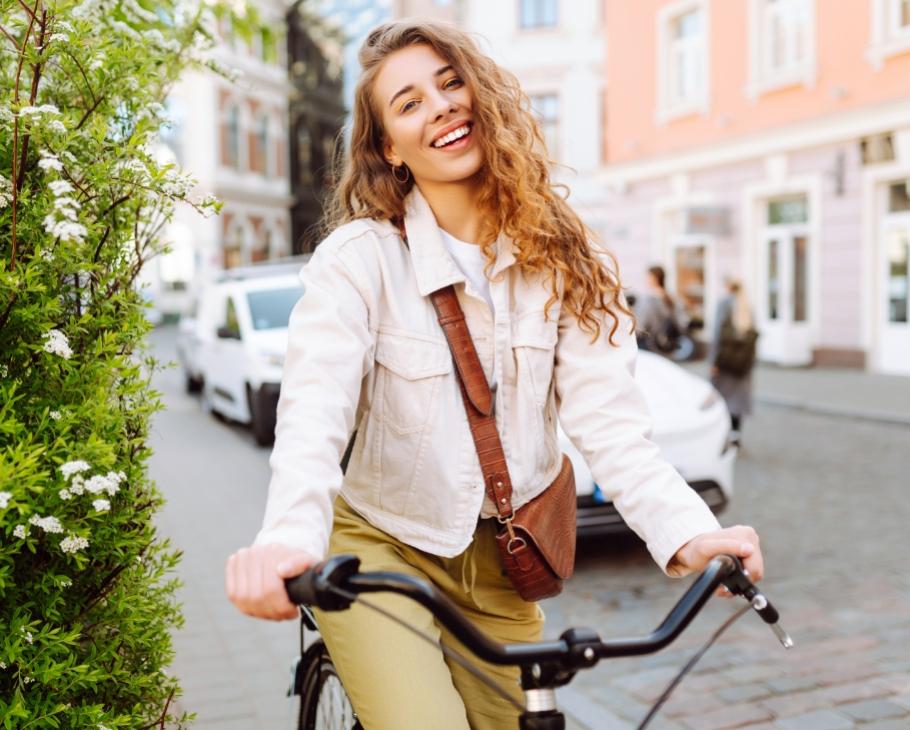 Donna sorridente in bicicletta su strada cittadina, indossa giacca chiara e borsa a tracolla.