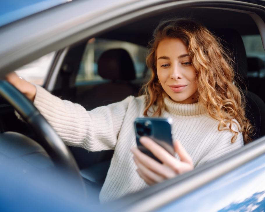 Donna in auto guarda il telefono, indossa un maglione bianco a collo alto.