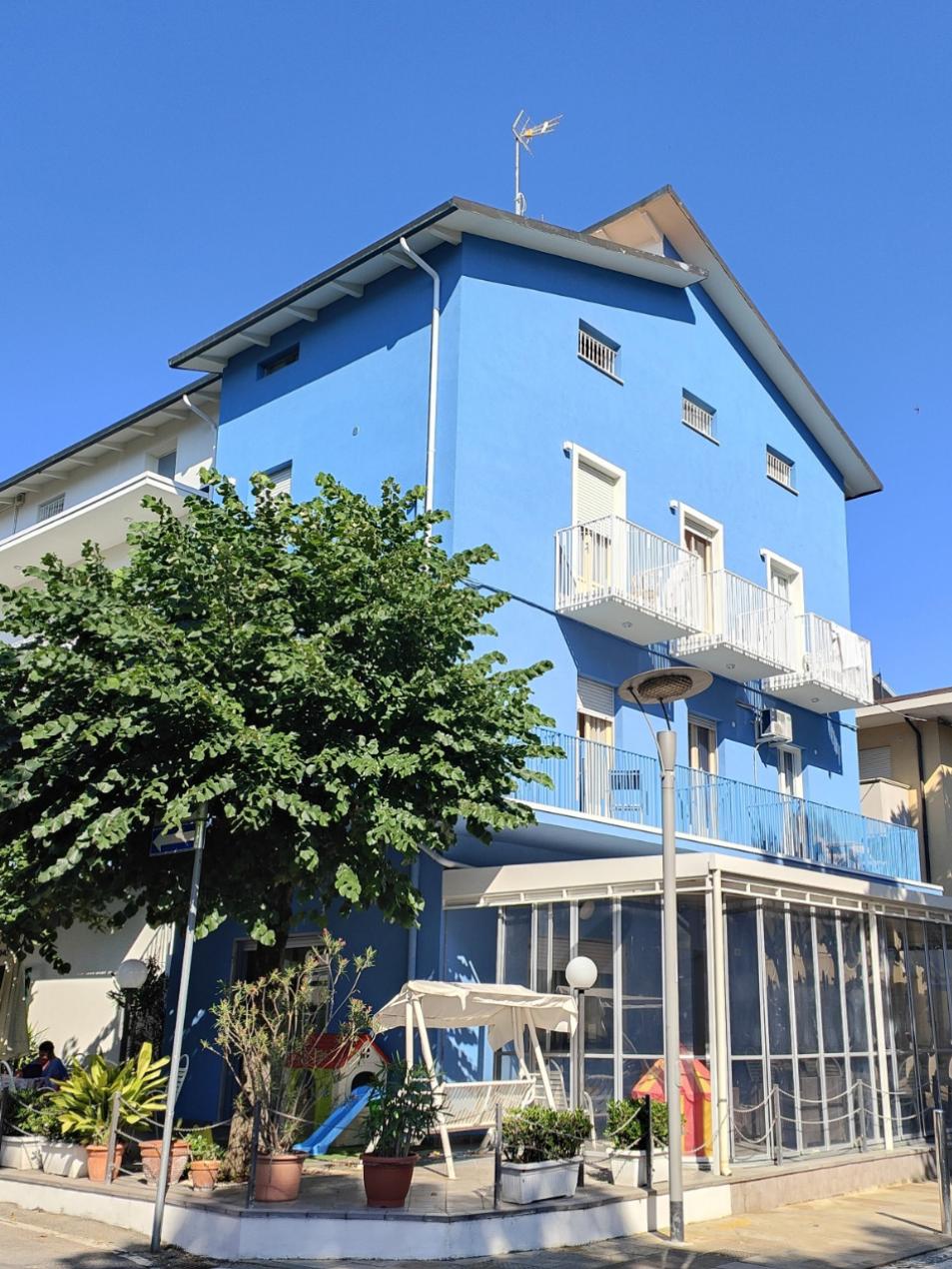 Edificio blu a tre piani con balconi bianchi e giardino con piante.